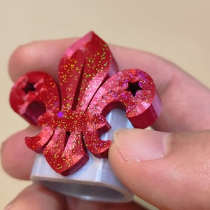 May include: A red plastic ring with a glitter finish featuring a fleur-de-lis design with three stars.