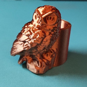 Puede incluir: Una figurita de búho de color cobre con detalles precisos, posada en una rama. El búho está mirando hacia la derecha y tiene las alas plegadas. La figurita es una pieza decorativa para el hogar o la oficina.