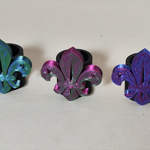 May include: Three 3D printed rings featuring a green, pink, and blue fleur-de-lis design.