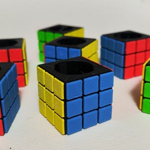 Puede incluir: Seis porta anillos en forma de cubo de diferentes colores con un agujero central negro. Cada cubo tiene una combinación de colores diferente de rojo, azul, amarillo y verde.