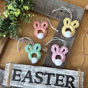 May include: Four handmade crochet bunny ornaments in pink, yellow, and green with white pom-pom tails. The ornaments are displayed on a rustic wooden frame with a sign that reads "EASTER". A green wreath and twine are also in the image.