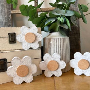 May include: White flower-shaped wooden knobs with a light brown center. The knobs are arranged on a wooden surface, with a vase of greenery and a decorative box in the background. The knobs are a decorative home decor item.