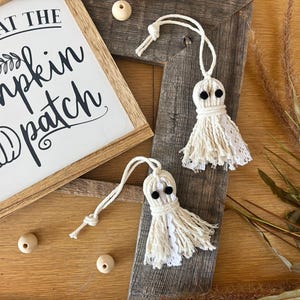 May include: Two handmade ghost ornaments with black button eyes and cream-colored yarn and lace details. A wooden sign with the text "Meet me at the pumpkin patch" is in the background. Wooden beads and dried grass complete the fall-themed arrangement.