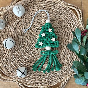 May include: A handmade green macrame Christmas tree ornament with white star accents and a tassel base. The ornament is displayed on a woven placemat with white bells and greenery, creating a festive holiday scene.
