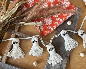 May include: A handmade Halloween garland featuring white yarn ghosts with black button eyes. The ghosts are strung together with white cord. The image also includes a fall-themed orange patterned fabric and wooden beads.
