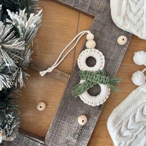 May include: A handmade macrame snowman Christmas ornament with a green scarf and wooden beads. The ornament is displayed on a wooden surface with other Christmas decorations, including a knitted stocking and a snow-covered pine branch.