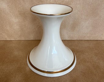 May include: A white ceramic candle holder with a flared, hourglass-shaped silhouette. The piece features a smooth, glossy finish and is accented with a thin, metallic gold trim along the top rim and the circular base, providing a clean look.