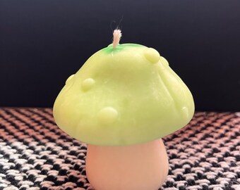 May include: A green and white mushroom shaped candle with a white wick. The candle is sitting on a black and white checkered surface.