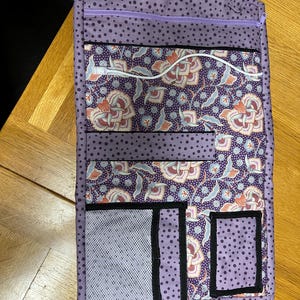 Könnte beinhalten: Lilafarbener Stofforganizer mit mehreren Taschen und einem Reißverschluss. Der Organizer hat ein Blumenmuster und schwarze Punkte. Er verfügt über eine Netztasche und zwei kleinere Taschen mit schwarzem Besatz.