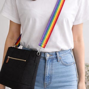 May include: Black crossbody bag with a rainbow strap. The bag has a gold zip and a small leather accent. The strap is a vibrant rainbow of colours. The person is wearing a white t-shirt and blue jeans.