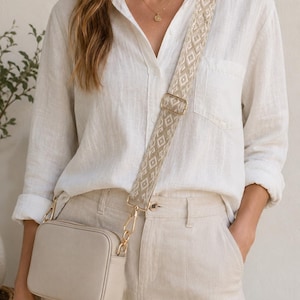 May include: A beige crossbody bag with a geometric patterned strap. The bag has a rectangular shape with gold-colored hardware. The strap is beige with a repeating diamond pattern. The bag is paired with a white button-down shirt and beige pants.