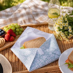 Könnte beinhalten: Eine Picknick-Szene mit einem Sandwich in einer wiederverwendbaren blauen Blumenverpackung. Eine klare Wasserflasche mit Zitronenscheiben, ein Strauß Gänseblümchen und ein Weidenkorb mit Snacks vervollständigen die Szene.