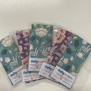Puede incluir: Cinco bolsas de tela con estampados florales en verde azulado, morado y rojo, cada una empaquetada en plástico transparente. Las bolsas tienen una etiqueta que dice "Crafting not Grafting" con información de contacto.