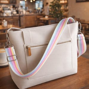 Könnte beinhalten: Cremefarbene Tragetasche mit einem regenbogenfarbenen Schultergurt. Die Tasche hat eine Reißverschlusstasche und einen braunen Leder-Reißverschlussanhänger. Die Tasche steht auf einem Holztisch in einem Café.