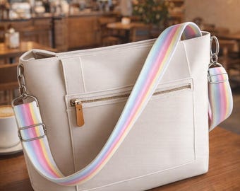 Puede incluir: Bolso tote color crema con una correa para el hombro a rayas arcoíris. El bolso tiene un bolsillo con cremallera y un tirador de cremallera de cuero marrón. El bolso está sobre una mesa de madera en un café.