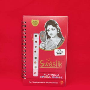 May include: A red spiral-bound booklet featuring a black and white illustration of a person holding a mirror. The cover displays the text Swastik, 576 ALL IN ONE MINI, and PLATINUM SPIRAL RANGE, with a vertical strip of small colorful bindi stickers.