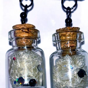 May include: A pair of dangle earrings featuring small glass bottles with cork stoppers. Each bottle is filled with iridescent glitter and white, translucent, shredded material.