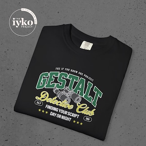 Puede incluir: Camiseta negra con un gráfico que dice "GESTALT Detective Club" en verde y amarillo. El diseño incluye binoculares y el texto "FINDING YOUR SCRIPT DAY OR NIGHT". La camiseta también presenta el logo "iyko PROJECT".