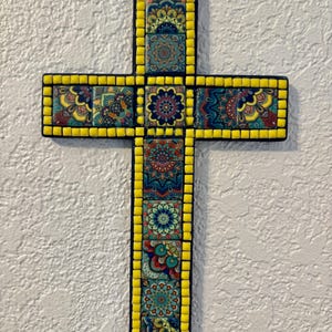 May include: A decorative cross with a mosaic design. The cross features a yellow border and is adorned with colorful square tiles displaying intricate floral patterns in shades of blue, red, and yellow. A small loop is attached for hanging.
