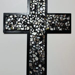 May include: A decorative cross with a black frame and a mosaic-style inlay of iridescent, irregularly shaped pieces. The mosaic has a mix of silver, gray, and black tones, creating a textured appearance. The cross is displayed against a white background.
