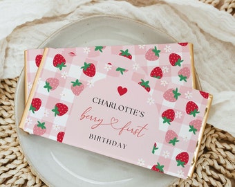 May include: A rectangular chocolate bar wrapper featuring a pink and white gingham pattern with red strawberry illustrations and small white flowers. The center displays a pink arch with the text CHARLOTTE'S berry first BIRTHDAY in black and red.
