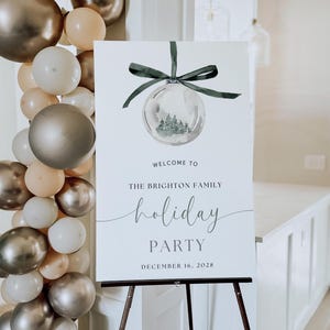May include: A white sign on a wooden easel with the text "Welcome to The Brighton Family Holiday Party December 16, 2028." The sign features a watercolor image of a Christmas ornament with a green bow. A balloon garland is on the left.