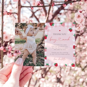 Berry First Birthday Thank You Card is shown printed horizontally as a 5x7" card. The left side has a space for a photo, and the right side includes an editable thank you note surrounded by red and pink strawberries, white daisies, and pink gingham. The top says thank you berry much.