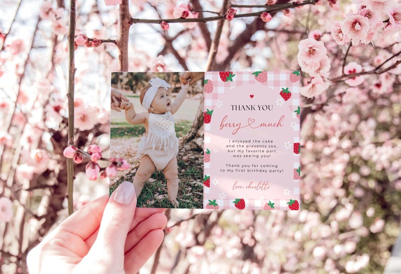 Berry First Birthday Thank You Card is shown printed horizontally as a 5x7" card. The left side has a space for a photo, and the right side includes an editable thank you note surrounded by red and pink strawberries, white daisies, and pink gingham. The top says thank you berry much.