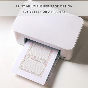 May include: A white printer with a sheet of paper emerging from the tray. The paper features two recipe cards printed on it, each with a colourful floral border. The text on the paper reads "Print Multiple Per Page Option (US Letter or A4 Paper)".