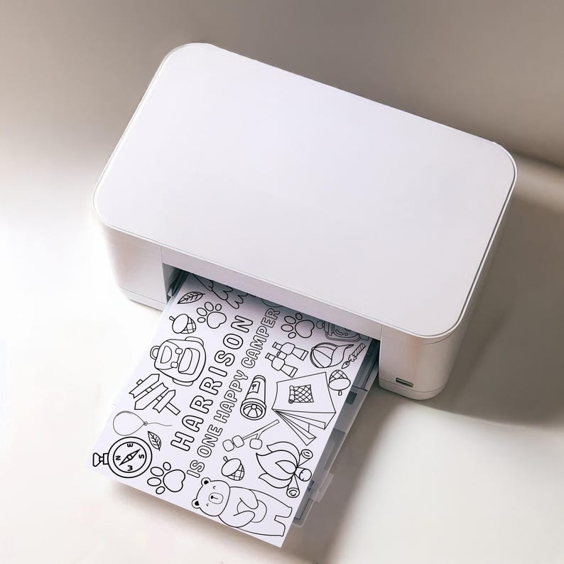 May include: A white printer with a sheet of paper coming out of the paper tray. The paper has a black and white illustration of a camping scene with the text "HARRISON IS ONE HAPPY CAMPER".