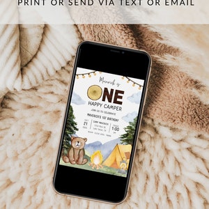May include: A digital invitation for a first birthday party with a woodland theme. The invitation features a cartoon bear, a campfire, a tent, and a wooden sign that says "Maverick is One Happy Camper". The invitation includes the date, time, and location of the party.