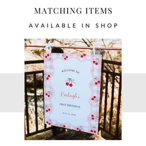 May include: A rectangular sign with a pink and white gingham border featuring red cherry illustrations. The text reads: MATCHING ITEMS AVAILABLE IN SHOP, WELCOME TO, Kaeleigh's, FIRST BIRTHDAY, JULY 21, 2029. The sign is suspended by white ribbons.