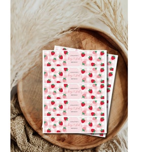 Puede incluir: Una pila de etiquetas de papel rectangulares con un patr&oacute;n de cuadros vichy rosa y blanco, ilustraciones de fresas rojas y flores blancas. El texto en cada etiqueta dice "CHALOTTE'S berry first BIRTHDAY" en fuente cursiva y sans-serif.