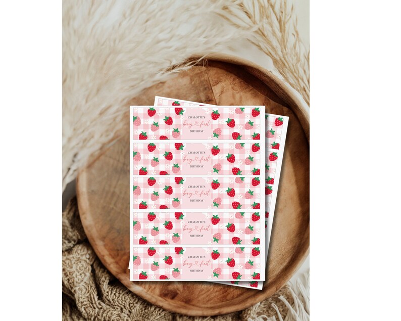 Puede incluir: Una pila de etiquetas de papel rectangulares con un patr&oacute;n de cuadros vichy rosa y blanco, ilustraciones de fresas rojas y flores blancas. El texto en cada etiqueta dice "CHALOTTE'S berry first BIRTHDAY" en fuente cursiva y sans-serif.