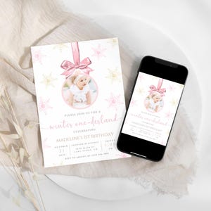 May include: A pink and white winter wonderland themed first birthday party invitation with a photo of a baby girl in a pink dress. The invitation features a pink and white snowflake ornament with a gold glitter background. The text on the invitation reads "Please join us for a winter one-derland celebrating Madeline's 1st Birthday".