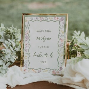 May include: A gold-framed sign with a white background and a green border. The sign says "Place your recipes for the bride-to-be here". The border is decorated with colourful drawings of fruits and vegetables.