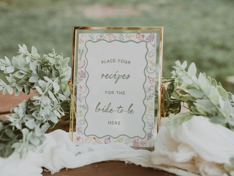 May include: A gold-framed sign with a white background and a green border. The sign says "Place your recipes for the bride-to-be here". The border is decorated with colourful drawings of fruits and vegetables.