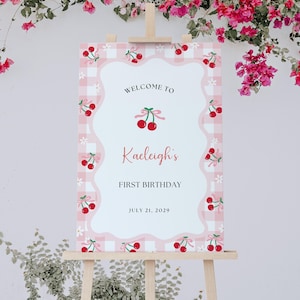 Puede incluir: Un letrero de bienvenida rosa y blanco a cuadros en un caballete de madera, decorado con cerezas y el texto "Welcome to Kaeligh's First Birthday July 21, 2029."