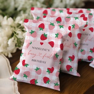 May include: A set of rectangular snack bags featuring a pink and white gingham pattern with red strawberry illustrations and small white flowers. A central label reads MADELINE'S berry first BIRTHDAY with a small red heart icon above the text.
