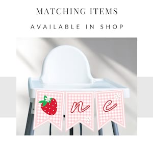 Op de afbeelding: Een roze en witte gingham-hoge stoelbanner met het woord "een" in rood. De banner heeft een aardbeienafbeelding aan het ene uiteinde en witte bloemen aan het andere.