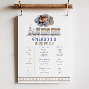 May include: A blue and white train themed birthday milestone poster with a photo of a young boy. The poster lists the boy's age, height, weight, favorite foods, songs, books, and things he can say and do. The poster says "Emerson's Second Birthday".