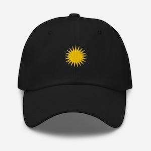 Könnte beinhalten: Schwarze Baseballkappe mit einem gelben, gestickten Sonnenmotiv auf der Vorderseite. Die Kappe hat einen gebogenen Schirm und einen verstellbaren Riemen auf der Rückseite. Das Sonnenmotiv ist zentriert und detailliert.