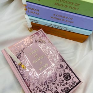 Pode incluir: Uma pilha de cinco livros de bolso com um livro rosa na frente. Os livros s&atilde;o todos parte da s&eacute;rie "A Court of Thorns and Roses" de Sarah J. Maas. Os livros est&atilde;o organizados em ordem de publica&ccedil;&atilde;o, com o livro mais novo no topo. O livro rosa &eacute; o primeiro livro da s&eacute;rie, "A Court of Thorns and Roses".