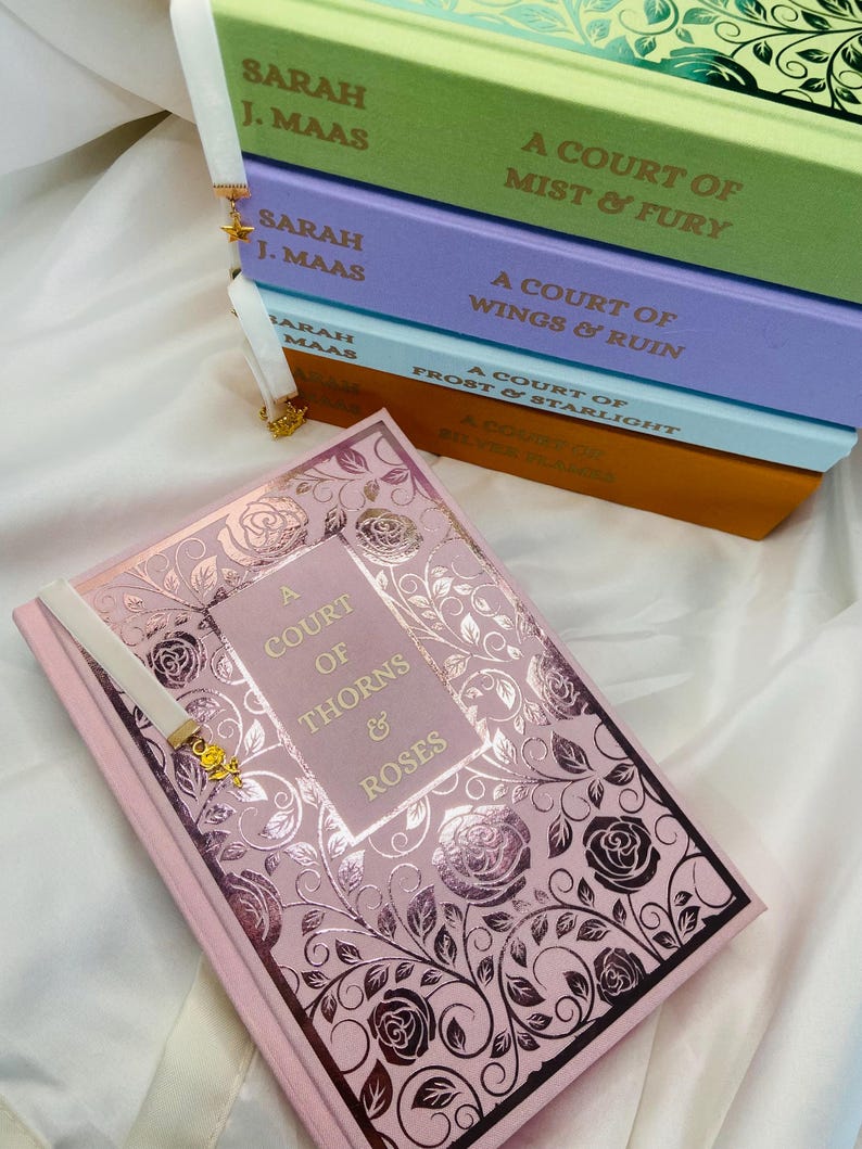 Pode incluir: Uma pilha de cinco livros de bolso com um livro rosa na frente. Os livros s&atilde;o todos parte da s&eacute;rie "A Court of Thorns and Roses" de Sarah J. Maas. Os livros est&atilde;o organizados em ordem de publica&ccedil;&atilde;o, com o livro mais novo no topo. O livro rosa &eacute; o primeiro livro da s&eacute;rie, "A Court of Thorns and Roses".