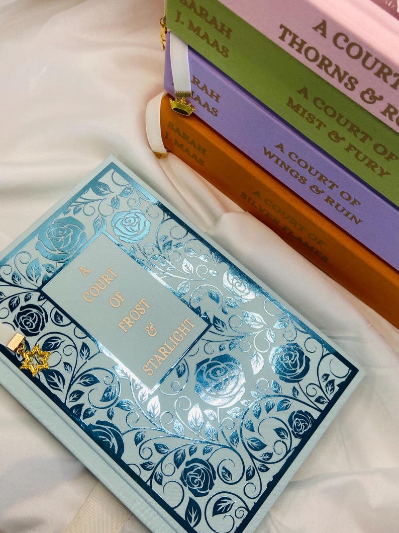 Pode incluir: Uma pilha de quatro livros de bolso com uma capa azul claro com um design floral prateado. Os livros s&atilde;o intitulados "A Court of Thorns & Roses", "A Court of Mist & Fury", "A Court of Wings & Ruin", e "A Court of Frost & Starlight".