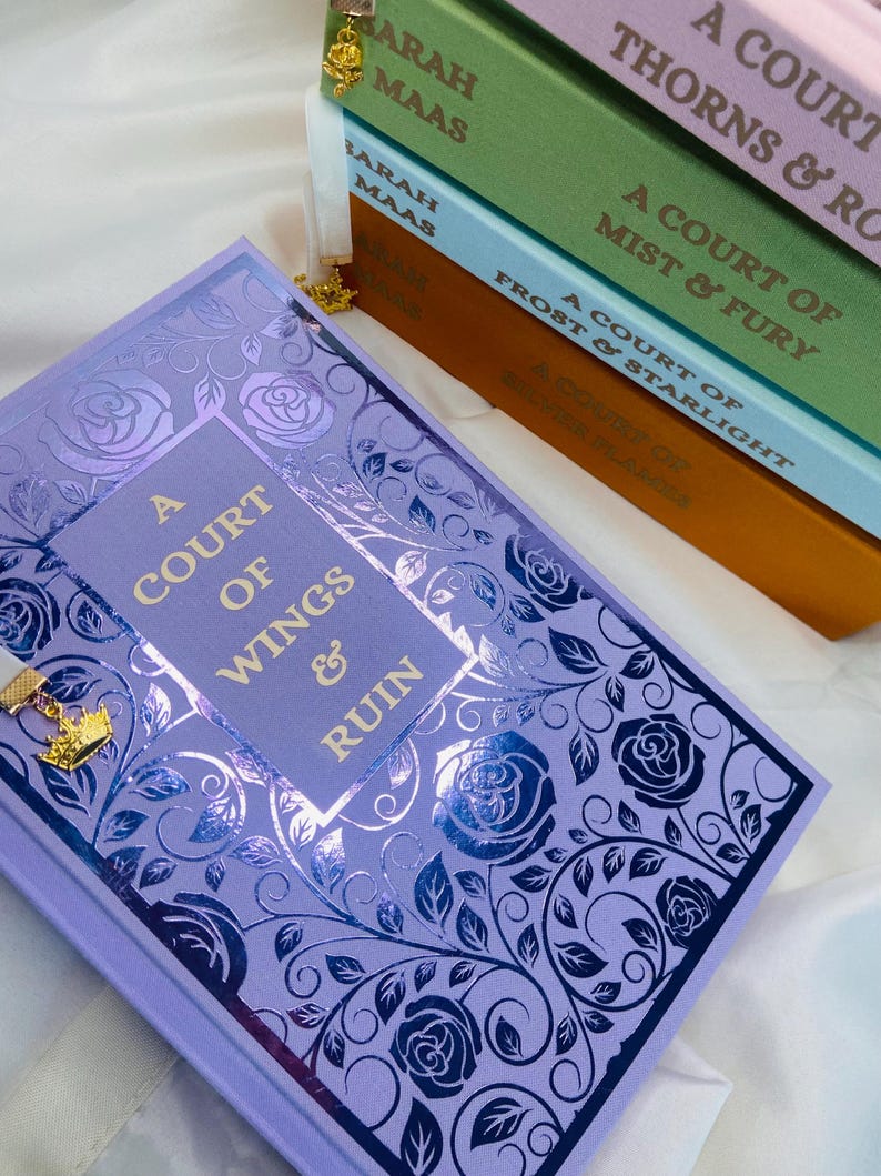Pode incluir: Um livro intitulado "A Court of Wings and Ruin" com uma capa roxa e um design floral prateado e preto. O livro faz parte de uma s&eacute;rie de Sarah J. Maas. O t&iacute;tulo do livro est&aacute; impresso em letras douradas.