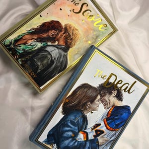 May include: Two hardback books titled The Score and The Deal by Elle Kennedy. The Score features a couple in a watercolour style with gold foil accents. The Deal shows a couple in a hockey jersey with gold foil lettering and a blue border.