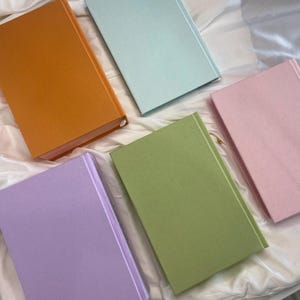 Pode incluir: Cinco cadernos de capa dura com diferentes cores de capa: laranja, azul claro, rosa, verde e lavanda.