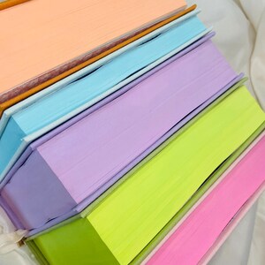 Pode incluir: Uma pilha de cinco livros com capas coloridas. As capas s&atilde;o p&ecirc;ssego, azul claro, lavanda, verde lim&atilde;o e rosa.