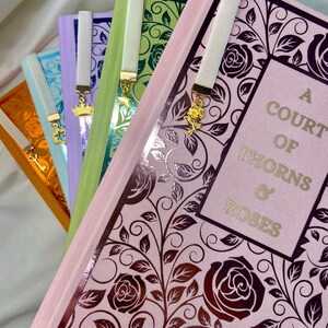 Pode incluir: Um conjunto de cinco livros com marcadores de fita de cetim combinando. Os livros s&atilde;o de cores diferentes e t&ecirc;m um design floral na capa. O livro de cima &eacute; rosa e tem o t&iacute;tulo "A Court of Thorns & Roses" na capa.
